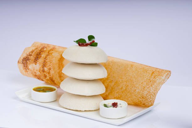 Ghee roast Dosa and Idlii, south indian main breakfast item which is beautifully arranged in a white plate lined with banana leaf and curry as sambar and chutney on white background.